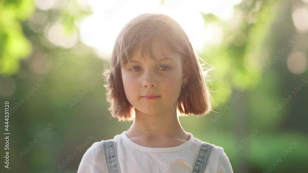 Beautiful little girl with bob haircut looking at camera and smiling in sunlight, her hair waving in wind. Child posing outside on sunny summer day, slow motion head and shoulders portrait