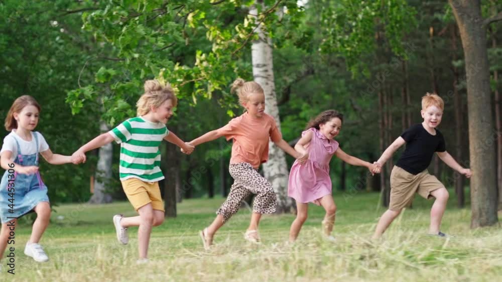 Group of five happy kids running on grass in green park holding hands and screaming with excitement. Children celebrating friendship and togetherness, side view slow motion shot
