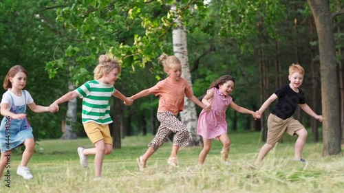 Group of five happy kids running on grass in green park holding hands and screaming with excitement. Children celebrating friendship and togetherness, side view slow motion shot
