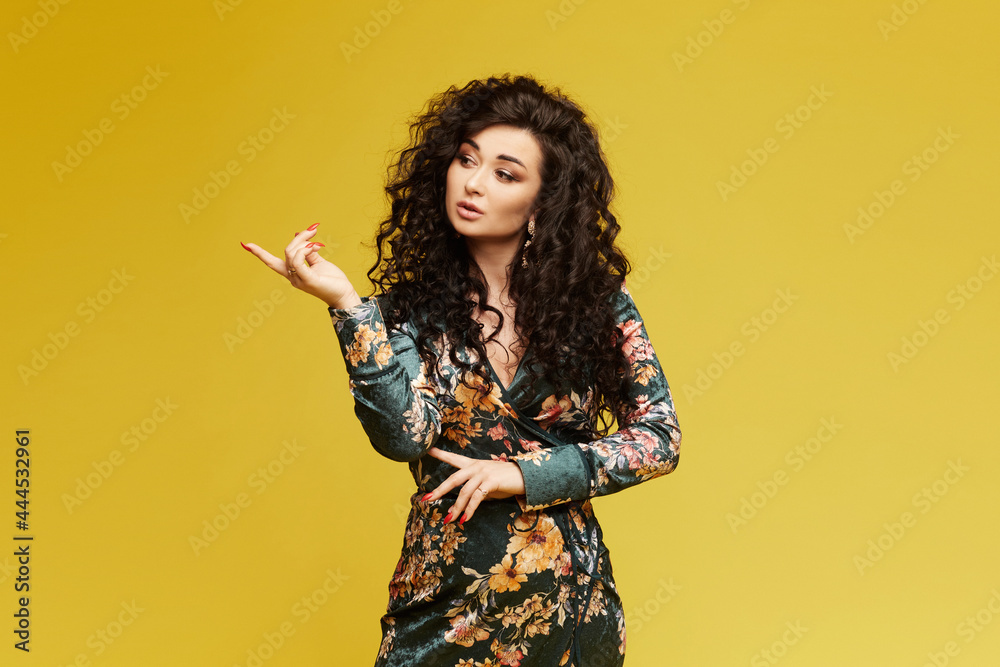 Fashion model girl with bright make-up in a trendy dress pointing finger on the left. A young brunette woman pointing finger on the left side of the shot isolated on yellow background Fashion model girl with bright make-up in a trendy dress pointing finger on the left. A young brunette woman pointing finger on the left side of the shot isolated on yellow background