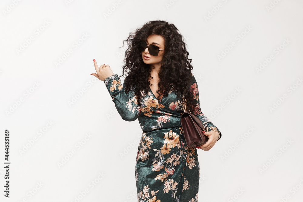 A young brunette woman with curly hair in trendy dress pointing her finger up and posing over white background. Model girl in fashionable clothes isolated on white background
