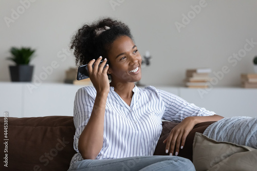 Excited African American woman listening to audio message, holding smartphone, sitting on cozy couch at home, smiling young female chatting online in social network by voice, using digital assistant