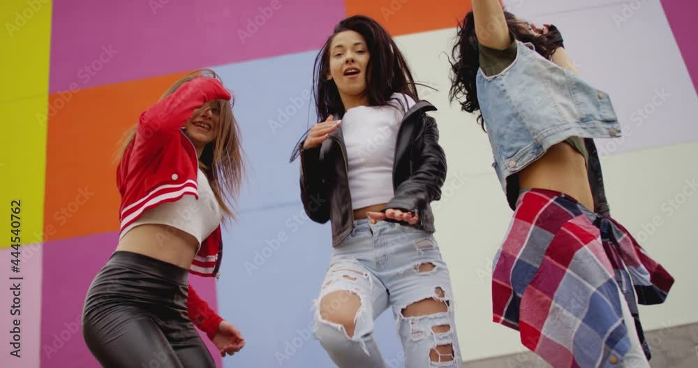 4K Three young beautiful , happy girls dancing , moving , looking at camera . Stylish bloggers in sport outfit having fun , jumping and shaking body in front of colorful background wall . slow motion 4K Three young beautiful , happy girls dancing , moving , looking at camera . Stylish bloggers in sport outfit having fun , jumping and shaking body in front of colorful background wall . slow motion