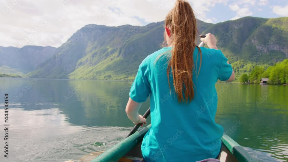 Handheld shot of a Caucasian young woman canoeing using a single-bladed paddle, traveling down a beautiful mountain lake, back view Handheld shot of a Caucasian young woman canoeing using a single-bladed paddle, traveling down a beautiful mountain lake, back view