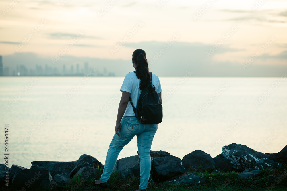woman with white sweater and black backpack standing watching the sea woman with white sweater and black backpack standing watching the sea
