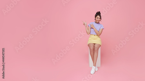 Happy beautiful Asian teen woman sitting on chair and hands pointing on pink background with copy space. panoramic background.