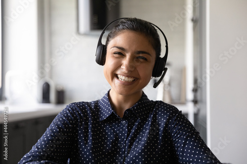 Happy funny Indian student wearing headphones and microphone looking at webcam, smiling at camera, laughing during virtual meeting or video call talk. Employee working from home. Screen view head shot