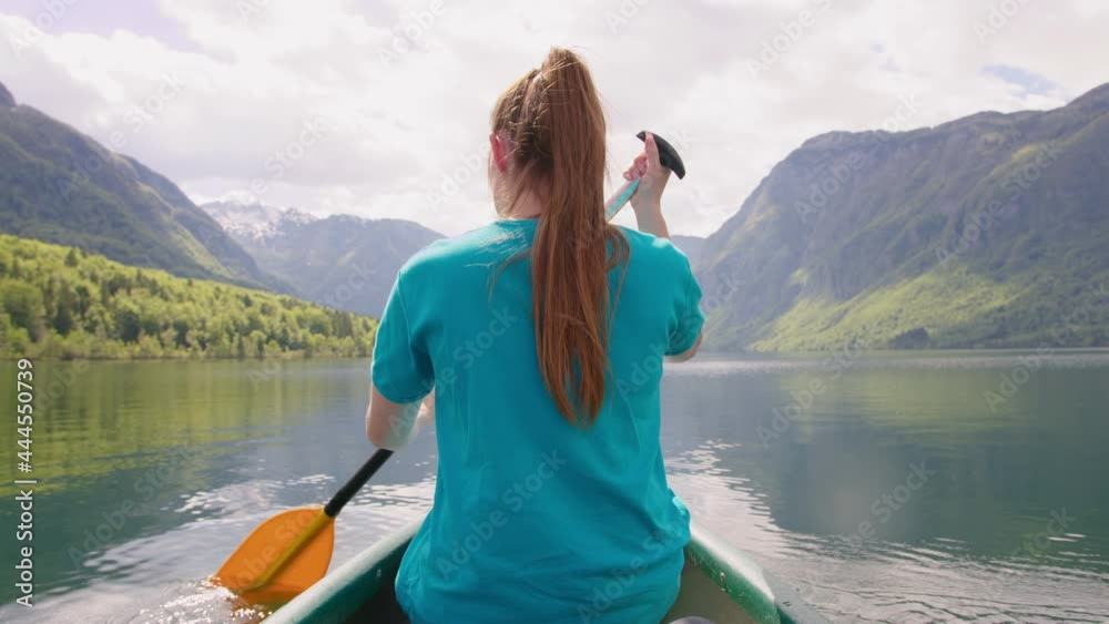 Handheld shot of a red-haired Caucasian girl steering the canoe in the direction of mountains, back view Handheld shot of a red-haired Caucasian girl steering the canoe in the direction of mountains, back view