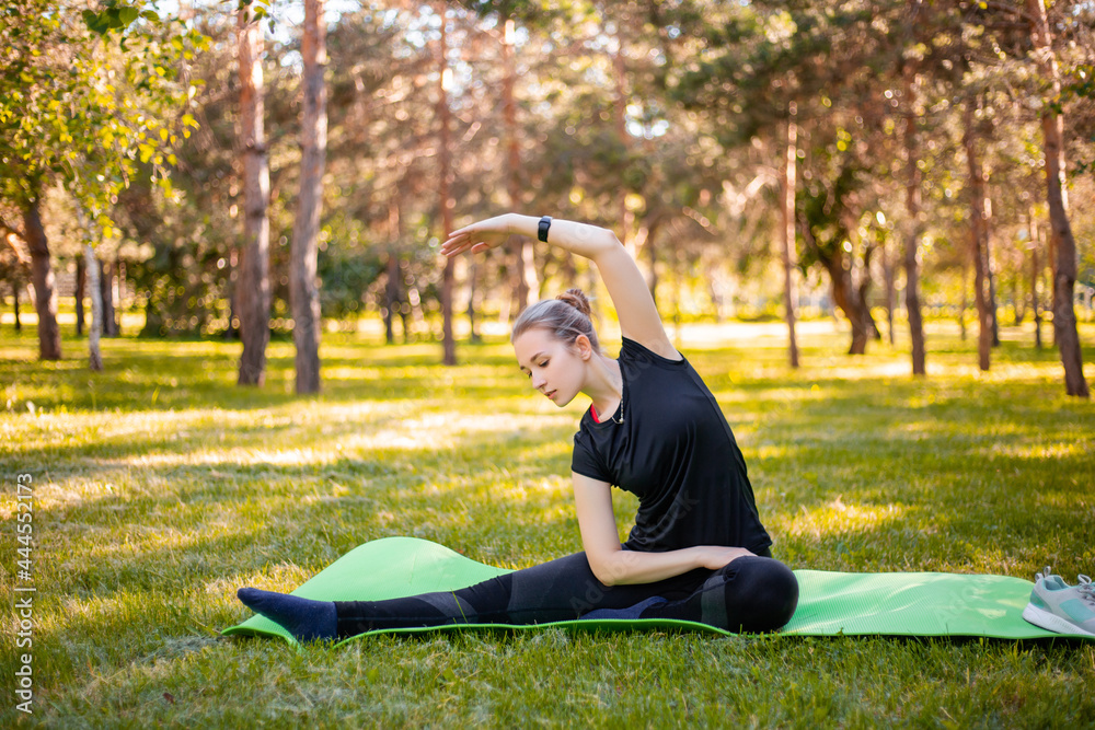 Outdoor Pilates training. A young woman does exercises to stretch the muscles of her legs while sitting on a yoga mat in the park. Exercise for the development of twine. Outdoor Pilates training. A young woman does exercises to stretch the muscles of her legs while sitting on a yoga mat in the park. Exercise for the development of twine.