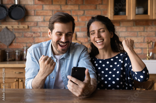 Excited millennial couple with smartphone celebrating success, approver loan, mortgage, getting good news, winning prize. Happy husband and wife using mobile phone, making winner yes gesture photo