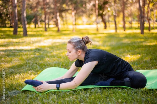 Outdoor Pilates training. A young woman does exercises to stretch the muscles of her legs while sitting on a yoga mat in the park. Exercise for the development of twine.