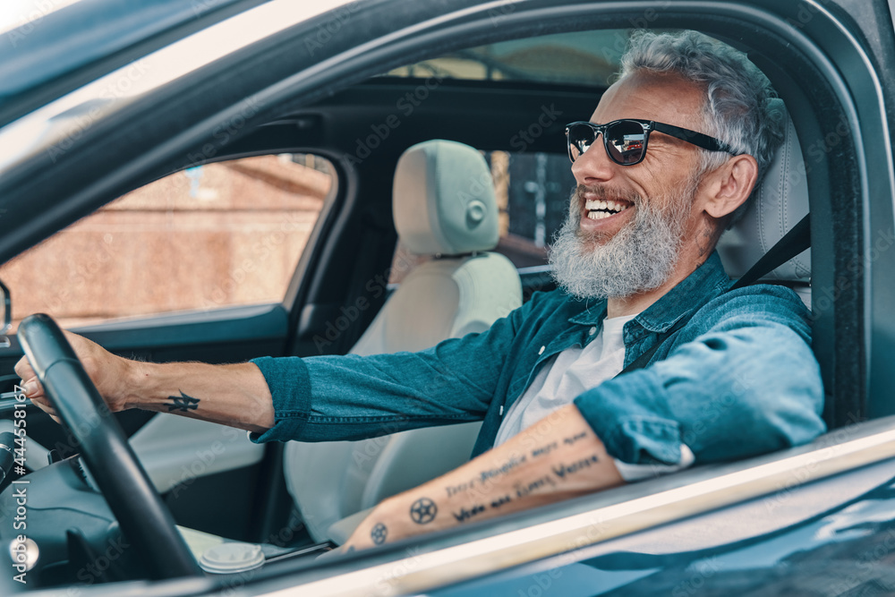 Carefree senior man in casual clothing enjoying car ride while sitting on front seat of the car