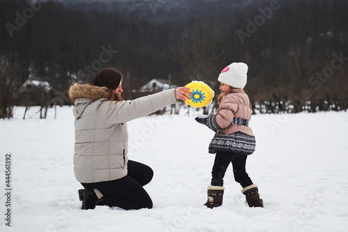 Young Caucasian pretty woman with long hair on walk with child during New Year and Christmas holidays. Joy of motherhood and winter fun. Mom and daughter play flying saucer in snow in winter park.