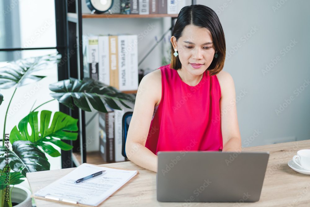 A cheerful middle-aged Asian businesswoman in relaxed casual dress working at home, checking email on computer laptop, writing on financial accounting document paper. Business stock photo