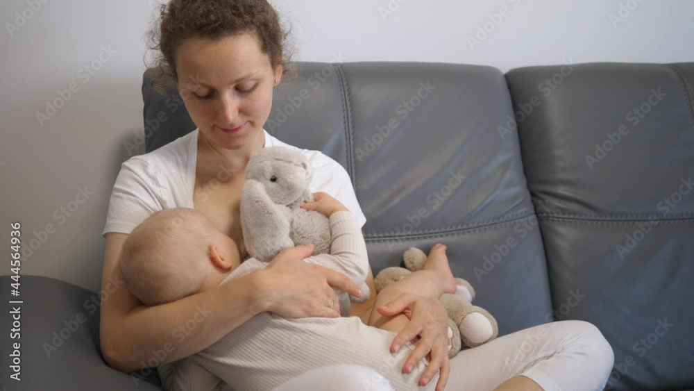 Young caucasian mom breastfeeds her baby on grey sofa. Baby is holding stuffed bunny Young caucasian mom breastfeeds her baby on grey sofa. Baby is holding stuffed bunny
