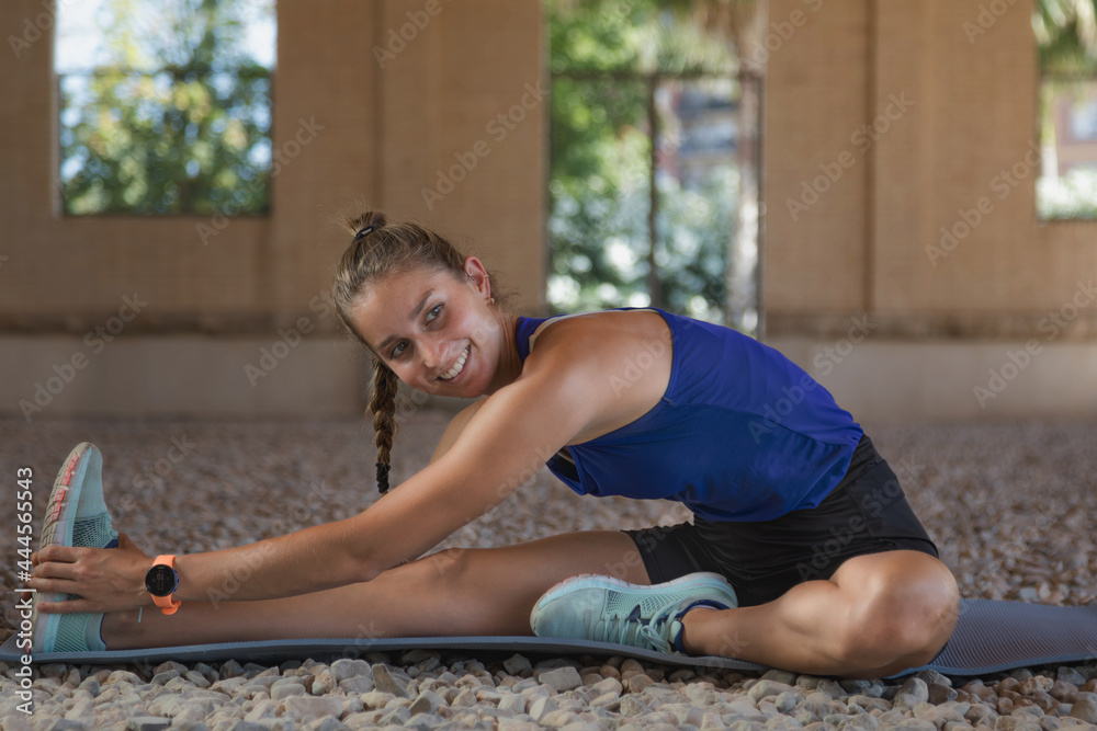 Smiling Caucasian woman in her 20s sitting on the floor doing stretching exercises looking camera