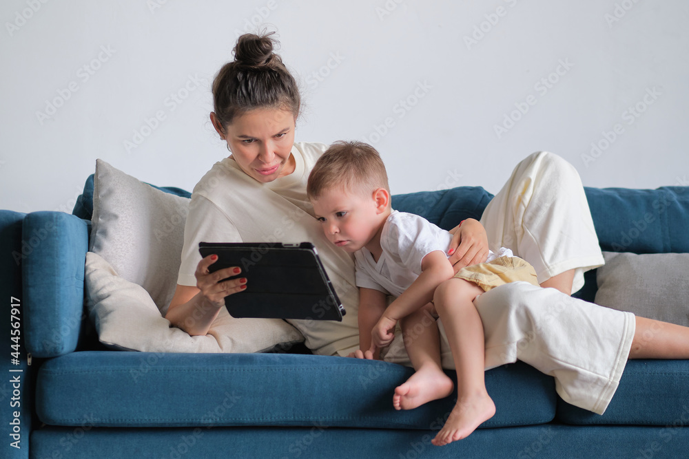 mother and son with tablet at home. Mother showing media content on line to her son in a tablet sitting on a couch in the living room in a house interior mother and son with tablet at home. Mother showing media content on line to her son in a tablet sitting on a couch in the living room in a house interior