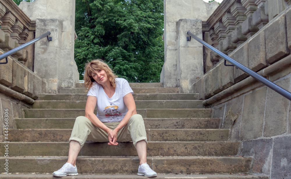 middle-aged woman, pretty blonde, sits on the steps in front of the park and smiles, female portrait in nature middle-aged woman, pretty blonde, sits on the steps in front of the park and smiles, female portrait in nature