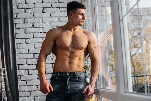 A young sexy athlete with perfect abs poses near the window in the studio topless in jeans on the background. Healthy lifestyle, proper nutrition, training programs and nutrition for weight loss.