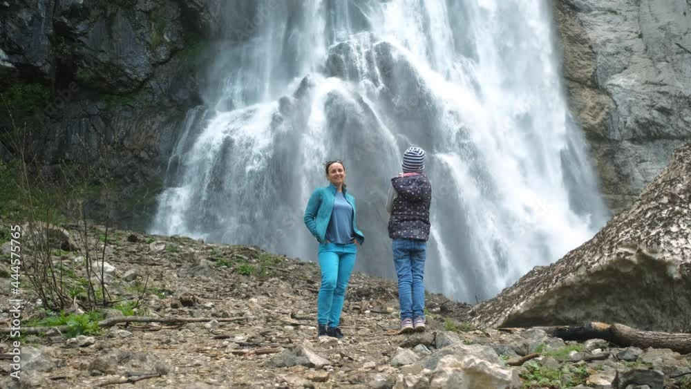 Cute Little Girl Taking Photos of her Mother by Beautiful Waterfall with the Smartphone, Slow Motion. Ecotourism and Travel Lifestyle Concept Cute Little Girl Taking Photos of her Mother by Beautiful Waterfall with the Smartphone, Slow Motion. Ecotourism and Travel Lifestyle Concept