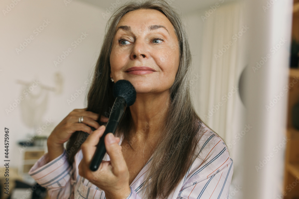 Smiling elderly woman doing makeup with brush in front of mirror at home. Attractive mature female looking with happiness. Personal care and aging concept Smiling elderly woman doing makeup with brush in front of mirror at home. Attractive mature female looking with happiness. Personal care and aging concept