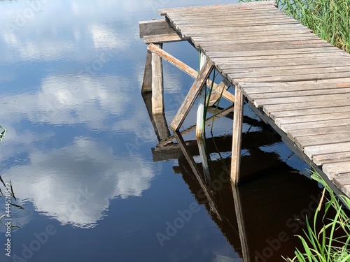 Wooden pier on the shore of the lake among the green reeds. Blue sky with white clouds. Summer landscape.