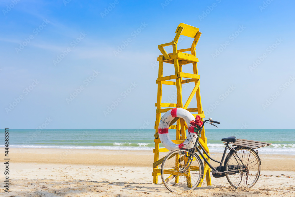 Fototapeta premium Old retro black bicycle with flowers bouquet beside Life ring for life safety on yellow lifeguard stand station or lifeguard chair protecting the safety of tourist on the empty beach in sunshine.