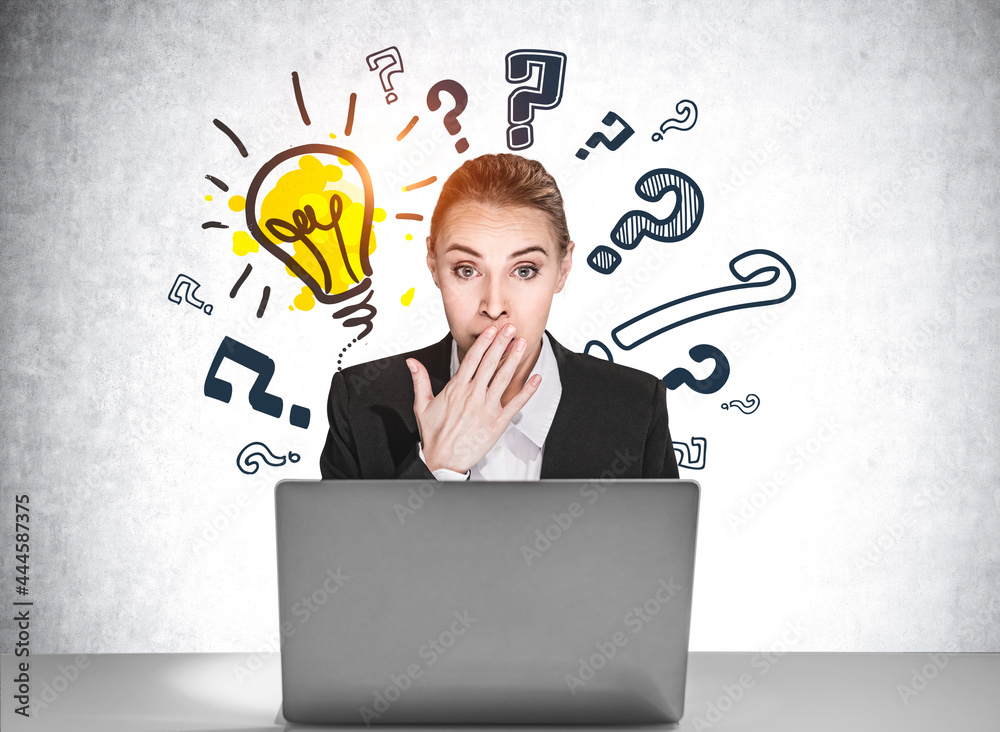 Portrait of amazed attractive business woman in formal wear sitting at the desk in front of laptop, sketches of question marks and light bulb drawn on concrete wall behind. Portrait of amazed attractive business woman in formal wear sitting at the desk in front of laptop, sketches of question marks and light bulb drawn on concrete wall behind.