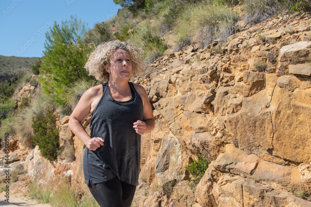 Naklejka premium curly haired blonde woman practicing running in nature on a sunny days
