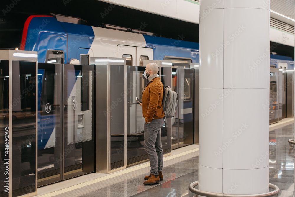 A man in a medical face mask to avoid the spread of coronavirus is holding a cellphone while waiting for a modern train on the subway. A bald guy in a surgical mask is keeping social distance. A man in a medical face mask to avoid the spread of coronavirus is holding a cellphone while waiting for a modern train on the subway. A bald guy in a surgical mask is keeping social distance.