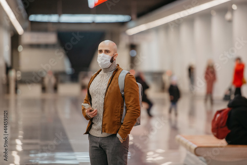 A man in a medical face mask is holding a smartphone and staring to the side while waiting for a train in the center of the subway station. A bald guy in a surgical mask is keeping social distance.