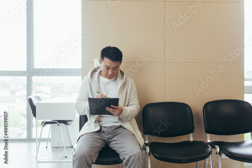 young asian male in waiting room for interview or meeting holding paper while sitting at chair fills out a resume questionnaire in office