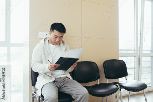 young asian male in waiting room for interview or meeting holding paper while sitting at chair fills out a resume questionnaire in office