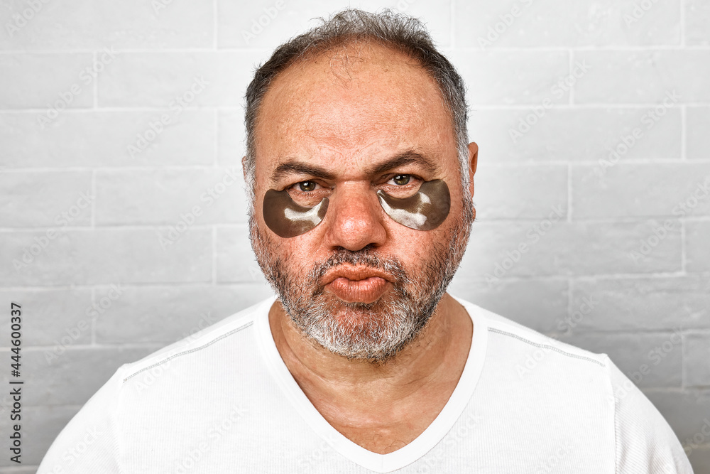 Obraz premium Handsome charming gray-haired bearded man with hydrogel eye patches on his face. Men's skincare morning routine.