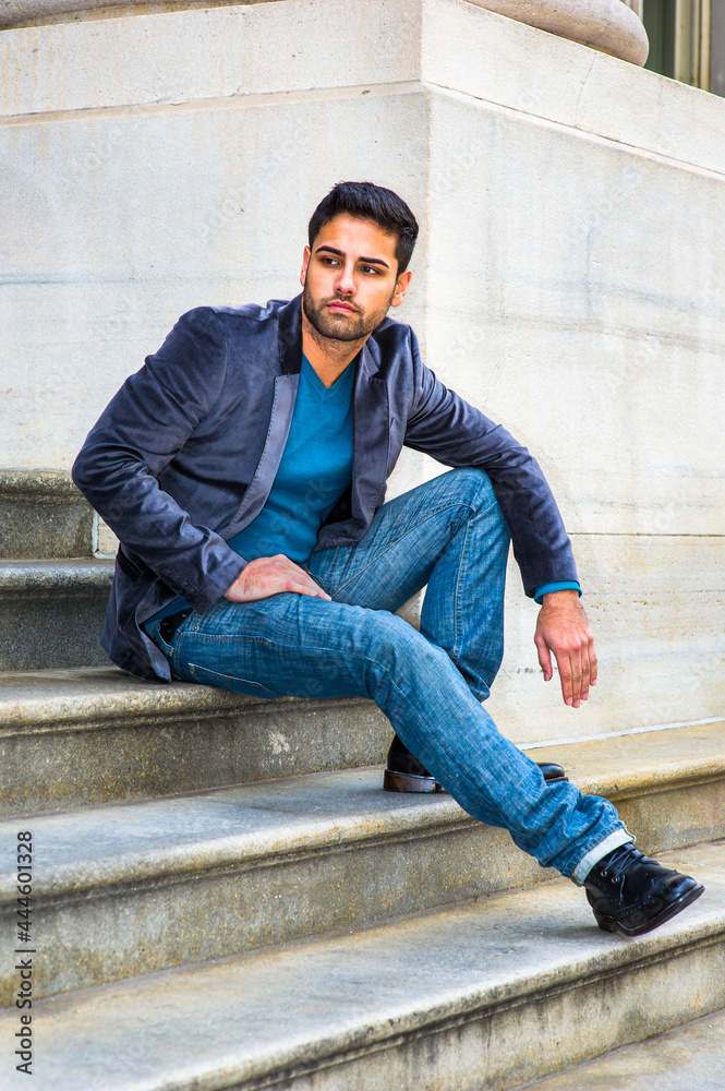 Dressing in a dark purple woolen blazer, blue jeans and black leather shoes, a young handsome Middle Eastern man is sitting on steps outside an office, thoughtfully looking around..