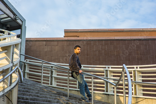Dressing in dark purple woolen blazer and jeans, a young guy with beard and mustache is standing against a metal railing on stairs, under sunshine of sunset, thinking.. © Alexander Image