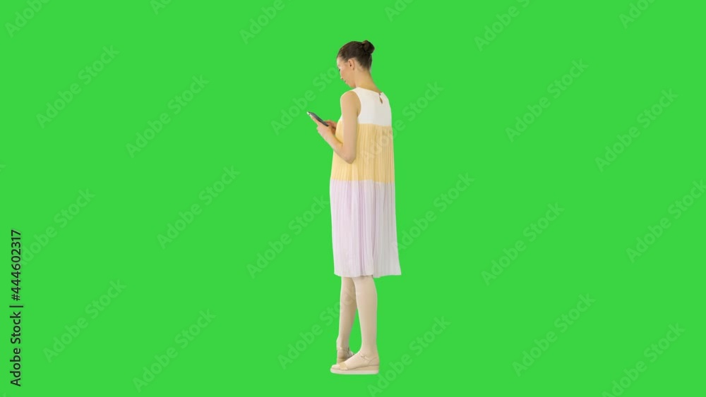 Young beautiful girl in white-yellow dress stands looking at her smartphone and smiles on a Green Screen, Chroma Key. Young beautiful girl in white-yellow dress stands looking at her smartphone and smiles on a Green Screen, Chroma Key.