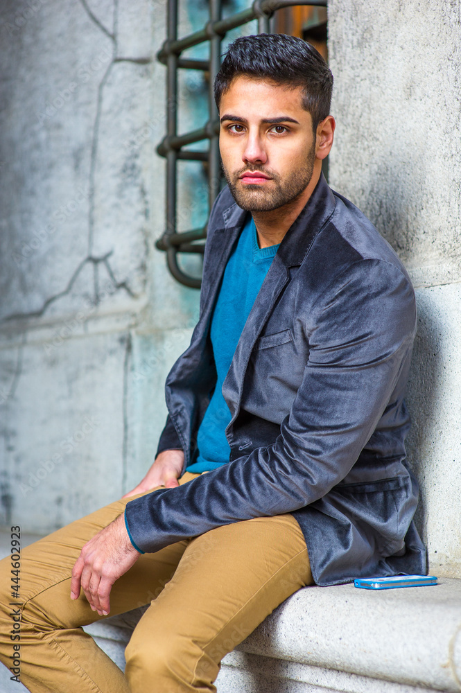 Dressing in a dark purple woolen blazer, a blue sweater and dark yellow pants, a young guy with bread and mustache is sitting outside, seriously looking at you..