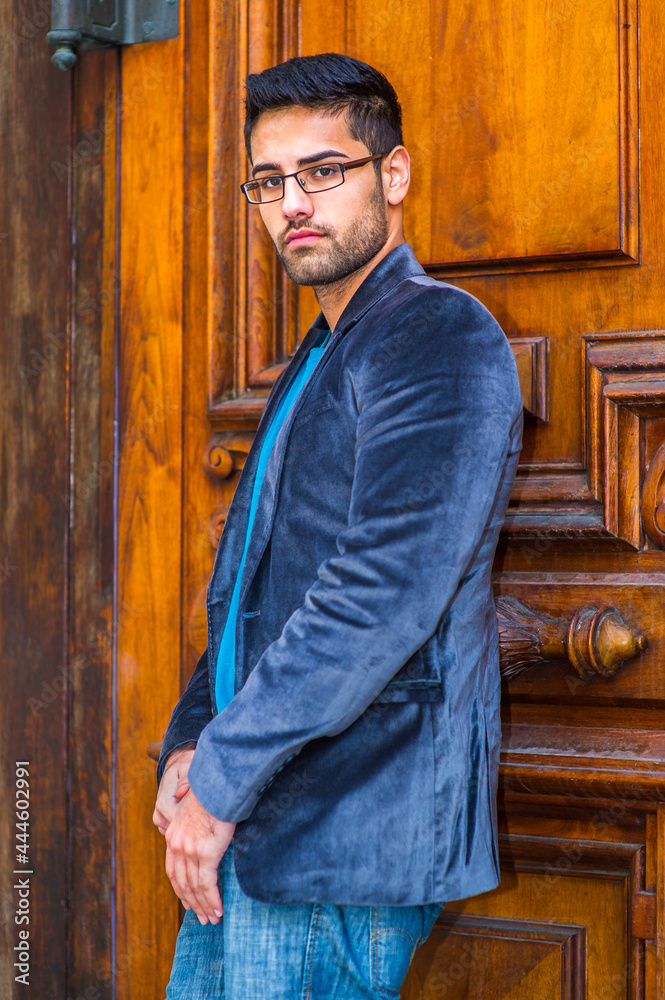 Dressing in a dark blue woolen blazer and jeans, wearing glasses, a young scholar with a little bread and mustache is standing in the front of door, thoughtfully looking at you.. Dressing in a dark blue woolen blazer and jeans, wearing glasses, a young scholar with a little bread and mustache is standing in the front of door, thoughtfully looking at you..