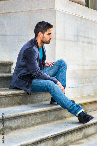 Dressing in a dark purple woolen blazer and blue jeans, a young handsome guy with a little bread and mustache is sitting on steps outside a office building, relaxing and thinking.