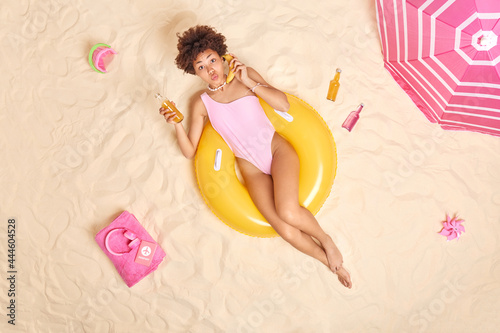 Summer time concept. Lovely dark skinned curly female wears bikini poses on inflated ring kees lips folded pretends having telephone talk holds banana near ear chills on beach during vacation photo