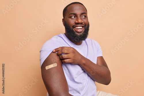 Health care vaccination and protection against covid 19 concept. Cheerful dark skinned bearded man shows shoulder with adhesive tape after being vaccinated received corona vaccine beige background