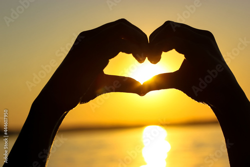 Silhouette of woman s hands in heart shape at sunset against a yellow-orange sky  sunbeams and a sun displayed inside. Symbol of love. Vacation by a sea  ocean  lake  river in summer. Seashore  coast.