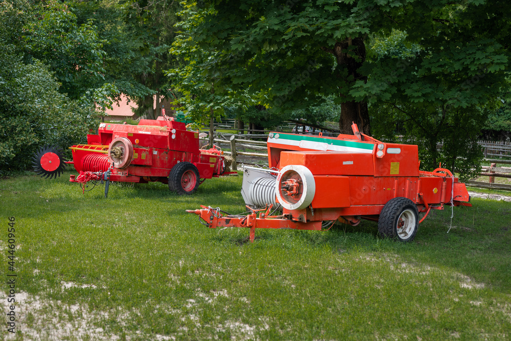 Colorful agricultural machinery - rural equipment standing on a farm, surrounded by trees on a sunny day Colorful agricultural machinery - rural equipment standing on a farm, surrounded by trees on a sunny day
