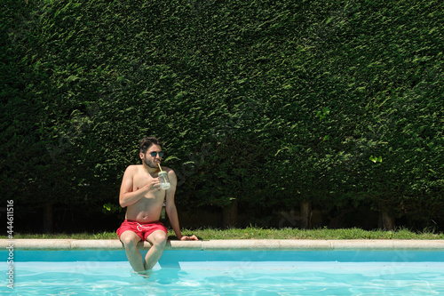 Young man wearing sunglasses drinking lemonade sitting on the swimming pool edge. Summer concept.