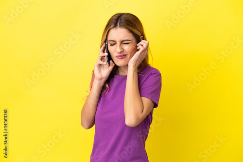 Young woman using mobile phone over isolated yellow background frustrated and covering ears © luismolinero