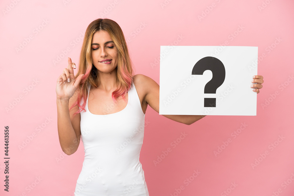 Young woman over isolated pink background holding a placard with question mark symbol with fingers crossing Young woman over isolated pink background holding a placard with question mark symbol with fingers crossing