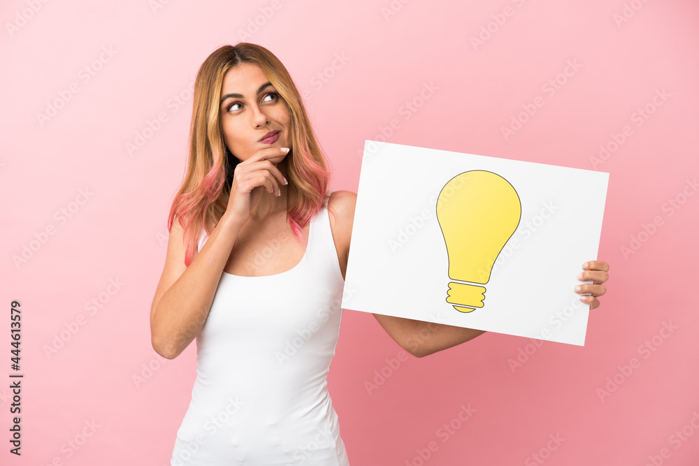 Young woman over isolated pink background holding a placard with bulb icon and thinking Young woman over isolated pink background holding a placard with bulb icon and thinking