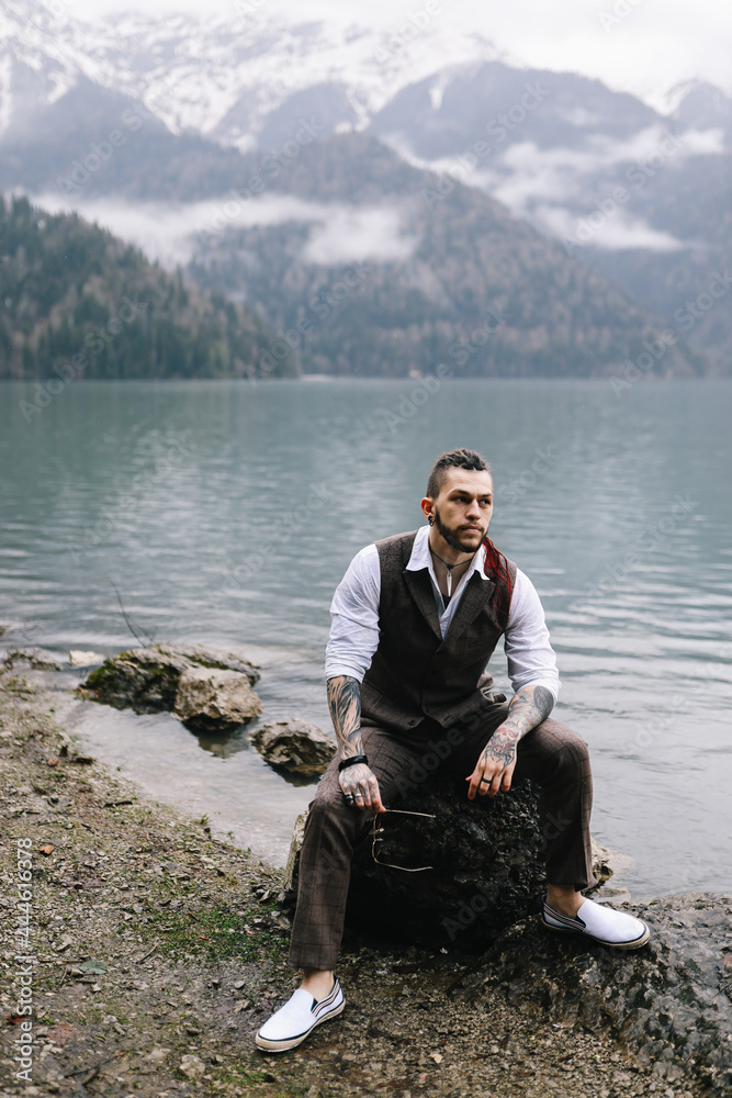 Fototapeta premium A brutal thoughtful young man groom with a dreadlocked hairstyle in a fashionable elegant wedding suit stands on the shore of a lake against the background of misty mountains in nature