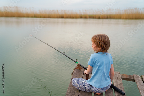 little girl sits on a wooden bridge on a river with fishing rod.